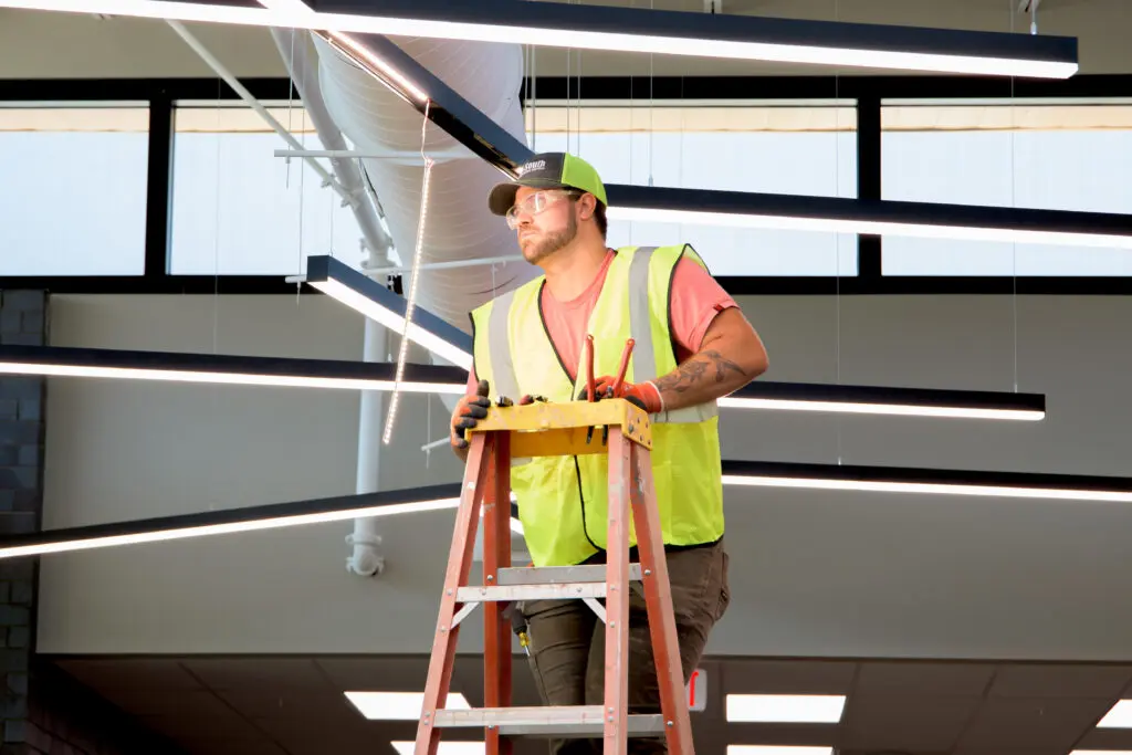 Electrician in hi-vis vest on tall orange ladder working on unique hanging lighting fixture