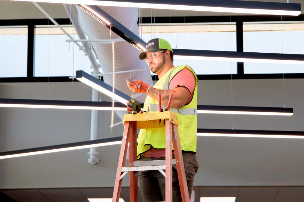 Electrician in hi-vis vest on tall orange ladder working on unique hanging lighting fixture